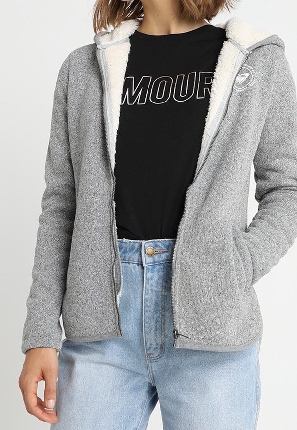 Femme portant une veste polaire gris clair zippée avec une doublure sherpa blanche sur un t-shirt noir avec le texte « AMOUR », assortie à un jean bleu clair.
