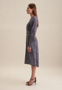 Robe bleu marine à manches longues avec un motif à pois blancs, corsage ajusté et jupe à hauteur de genoux. Portée avec des sandales à talons hauts noires.