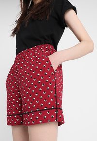 Röda shorts med ett geometriskt svartvitt mönster, elastisk midjeband och fickor; avslutade med en svart kant längs fållen.