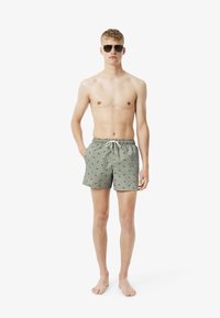 Groene zwembroek met een trekkoord in de taille, voorzien van een patroon van kleine groene bladeren, gecombineerd met een zonnebril, het model staat blootsvoets.