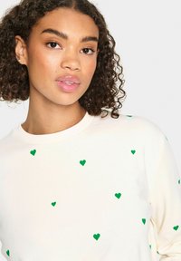 Sudadera de color crema claro, hecha de una tela suave, con un diseño de corazones verdes dispersos. El cuello es redondo y las mangas cuentan con puños acanalados que completan el diseño.