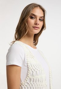 Gilet all'uncinetto in filato off-white con motivi geometrici e bordi frangiati, indossato sopra una maglietta bianca semplice. Texture morbida e design aperto.