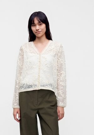 Jonge vrouw met lang donker haar draagt een doorschijnende witte blouse met bloemenkant en lange mouwen en donkergroene broek met hoge taille.