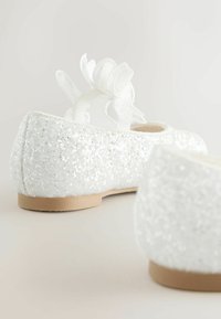 Balerini albi cu suprafață sclipitoare, având un accent floral pe o curea transparentă și o talpă de cauciuc bej.