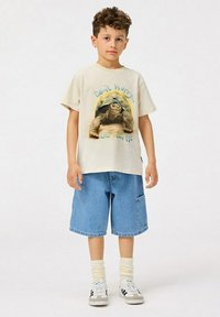 Beige puuvillane T-särk, millel on kilpkonna graafika ja tekst, koos helesiniste denim lühkaritega. Valged ja hallid spordijalatsid, mis on ühendatud kreemikat värvi sokkidega.