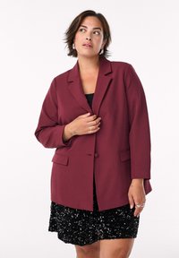 Blazer borgogna con maniche lunghe, rever a punte e due tasche frontali, abbinato a una gonna nera di paillettes. Texture liscia.