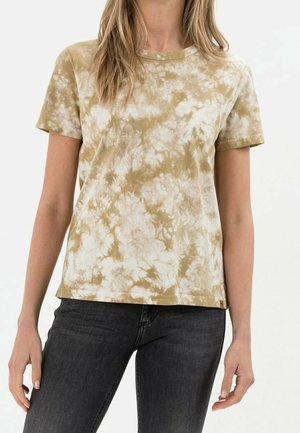 Femme portant un t-shirt à manches courtes beige et blanc tie-dye associé à un jean gris foncé sur un fond uni.