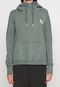 Felpa verde con cappuccio, tasca frontale, maniche lunghe, lacci e un piccolo logo bianco sul petto sinistro, indossata da una persona.