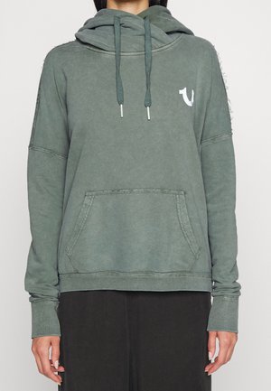 Felpa verde con cappuccio, tasca frontale, maniche lunghe, lacci e un piccolo logo bianco sul petto sinistro, indossata da una persona.