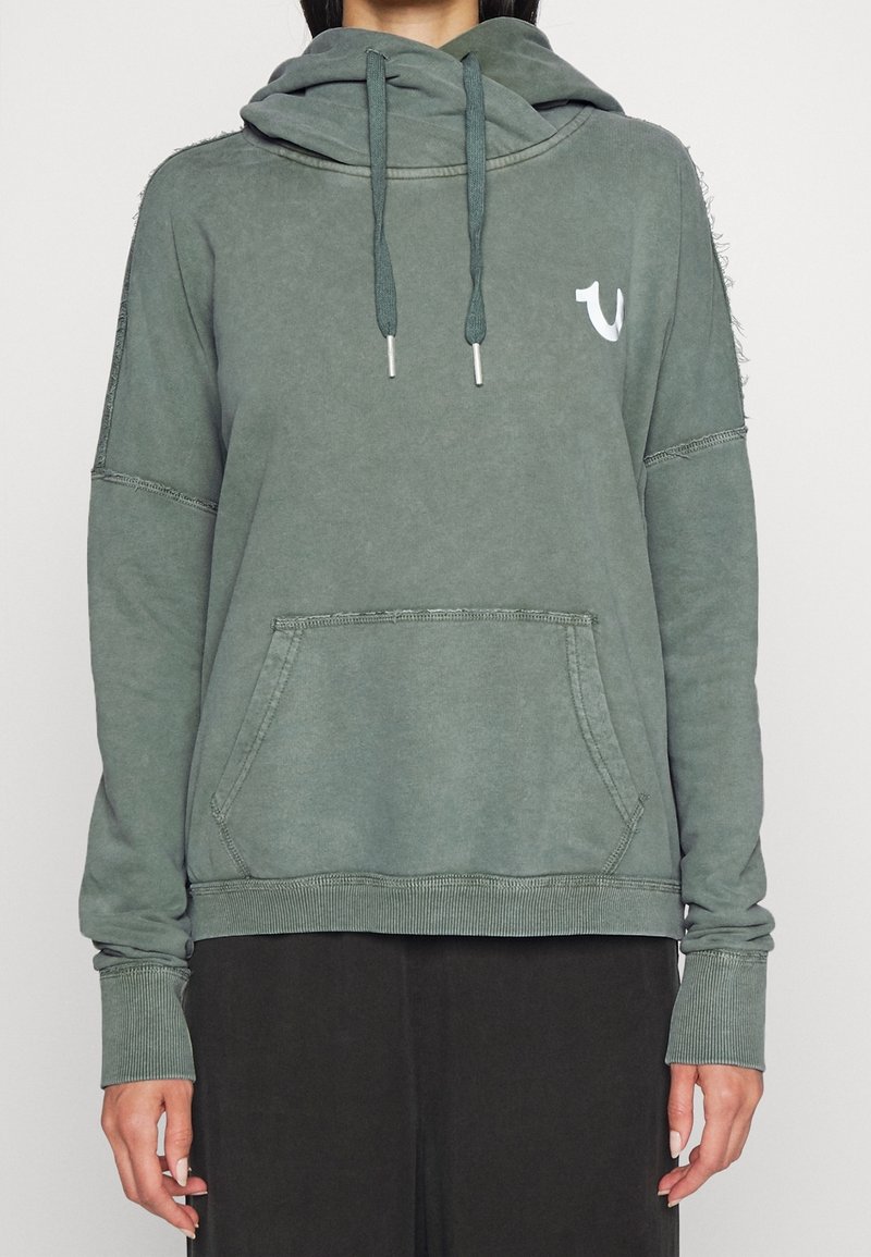 Felpa verde con cappuccio, tasca frontale, maniche lunghe, lacci e un piccolo logo bianco sul petto sinistro, indossata da una persona.