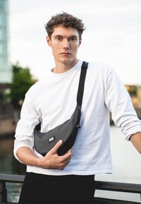 Čierna taška cez rameno vyrobená z hladkého materiálu, s oblým tvarom a výraznou značkovou etiketou. Model má na sebe biele tričko s dlhým rukávom.