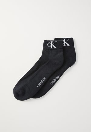 Calcetines de algodón negros con puños acanalados, que presentan el logo en blanco "CK" y el texto "Calvin Klein" en el costado. Textura suave y diseño de longitud a los tobillos.
