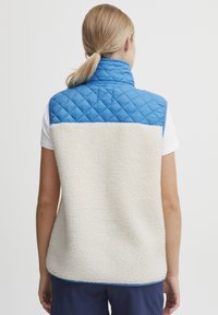 Blå quiltet øvre del med høy krage, kremfarget teksturert fleece i nedre del, ermeløs design og blå dekor langs kanten.