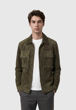 Jonge man met kort bruin haar draagt een olijfgroene suède jas over een wit t-shirt en grijze broek, staand met handen in de zakken.