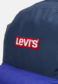 Sac à dos Levi's en tissu texturé bleu marine, avec un logo rouge brodé en évidence et une section inférieure violette contrastante.