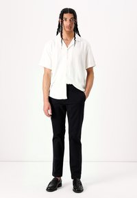 Camisa blanca de manga corta, tipo botón y de corte relajado, combinada con pantalones negros y zapatos negros pulidos. Diseño sencillo y de color sólido.