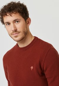 Maglione lavorato a maglia color bordeaux con colletto a girocollo, caratterizzato da un sottile emblema ad albero sul petto. Morbido al tatto e vestibilità classica.