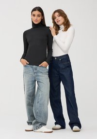 Dwie kobiety noszą dopasowane golfy – jeden czarny, drugi biały. Obie łączą je z luźnymi dżinsami i białymi trampkami. Neutralne tło.