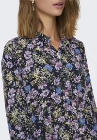Camicia floreale con base blu scuro, caratterizzata da fiori viola, blu e verdi. Presenta un collo a camicia e un design plissettato.