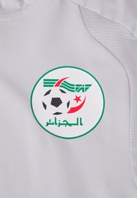 Λευκό ύφασμα με κυκλικό λογότυπο που απεικονίζει μια μαύρη μπάλα ποδοσφαίρου, πράσινες λεπτομέρειες και αραβικά κείμενα. Η σχεδίαση περιλαμβάνει μια κόκκινη ημισέληνο και ρίγες.