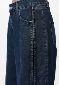 Mörkblå denimjeans har en avslappnad passform, vida ben och synliga sidosemsor. De har två framfickor och en enda bakficka.