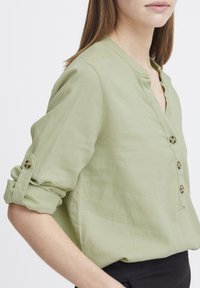 Chemise vert clair à boutons, en tissu doux. Présente un col en V, des manches roulées avec des accents de boutons, et une coupe décontractée. Design simple.