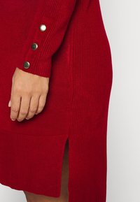 Robe en maille côtelée rouge avec une fente à l'ourlet et des boutons dorés décoratifs aux poignets, mettant en valeur la texture et les détails de conception.