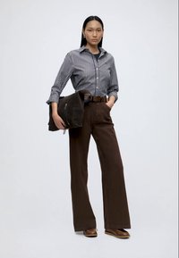 Femme portant une chemise rayée boutonnée, un pantalon large marron, des chaussures marron, tenant un sac marron foncé avec une main dans la poche.
