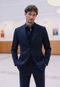Costume marine en tissu lisse, avec un design à un bouton et des revers en pointe. Porté avec une chemise sombre assortie et un pantalon droit.