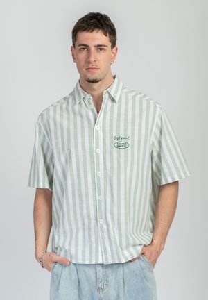 Ung mann iført en grønn og hvit kortermet skjorte med vertikale striper og tekst på brystet, hendene i lommene på lyse denimjeans.