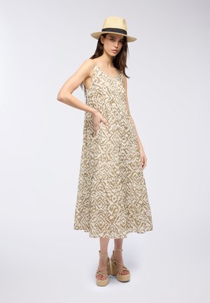 Abito maxi beige a fantasia con silhouette svasata, tasche laterali e spalline sottili. Il modello indossa un cappello di paglia e sandali con zeppa.