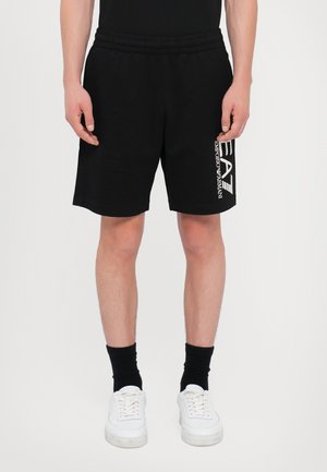 Osoba ubrana w czarne szorty Emporio Armani EA7, czarne skarpetki i białe trampki, stojąca na jednolitym tle.