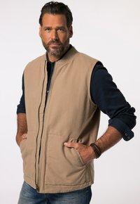 Beige ärmellose Weste mit einem Reißverschluss auf der Vorderseite, zwei Fronttaschen und einem gerippten Kragen, die über einem dunklen Shirt getragen wird und einen lockeren Sitz hat.