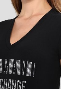 Zbliżenie na osobę z czarną koszulką w serek, na której częściowo widoczny jest biały tekst w kropki.