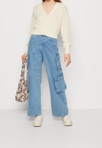 Krämbärgad stickad kofta med V-hals och knappstängning, kombinerad med lösa blå cargobyxor med sidofickor och ljusbruna plattformsskor.