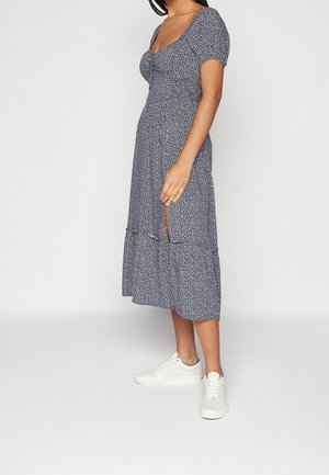 Femme portant une robe midi à fleurs bleues avec manches courtes, une fente latérale et des baskets blanches, posant devant un fond uni.