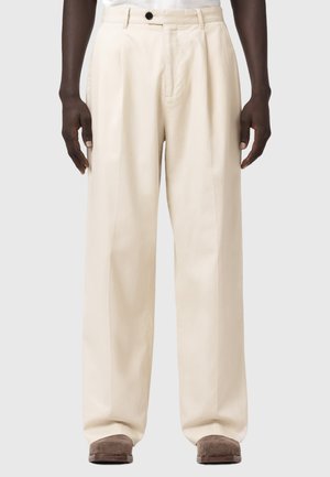 Osoba o ciemnej karnacji, ubrana w luźne, kremowe spodnie i brązowe buty, stojąca z rozluźnionymi ramionami wzdłuż ciała na jednolitym tle.