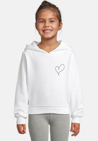Smilende jente i hvit hettegenser med et lite svart hjerte på brystet og grå leggings foran en enkel bakgrunn.
