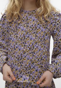 Fialové květinové šaty s žlutými a černými akcenty; řasená textura, dlouhé rukávy a elastické manžety. Hladká tkanina s jemným vzorem.