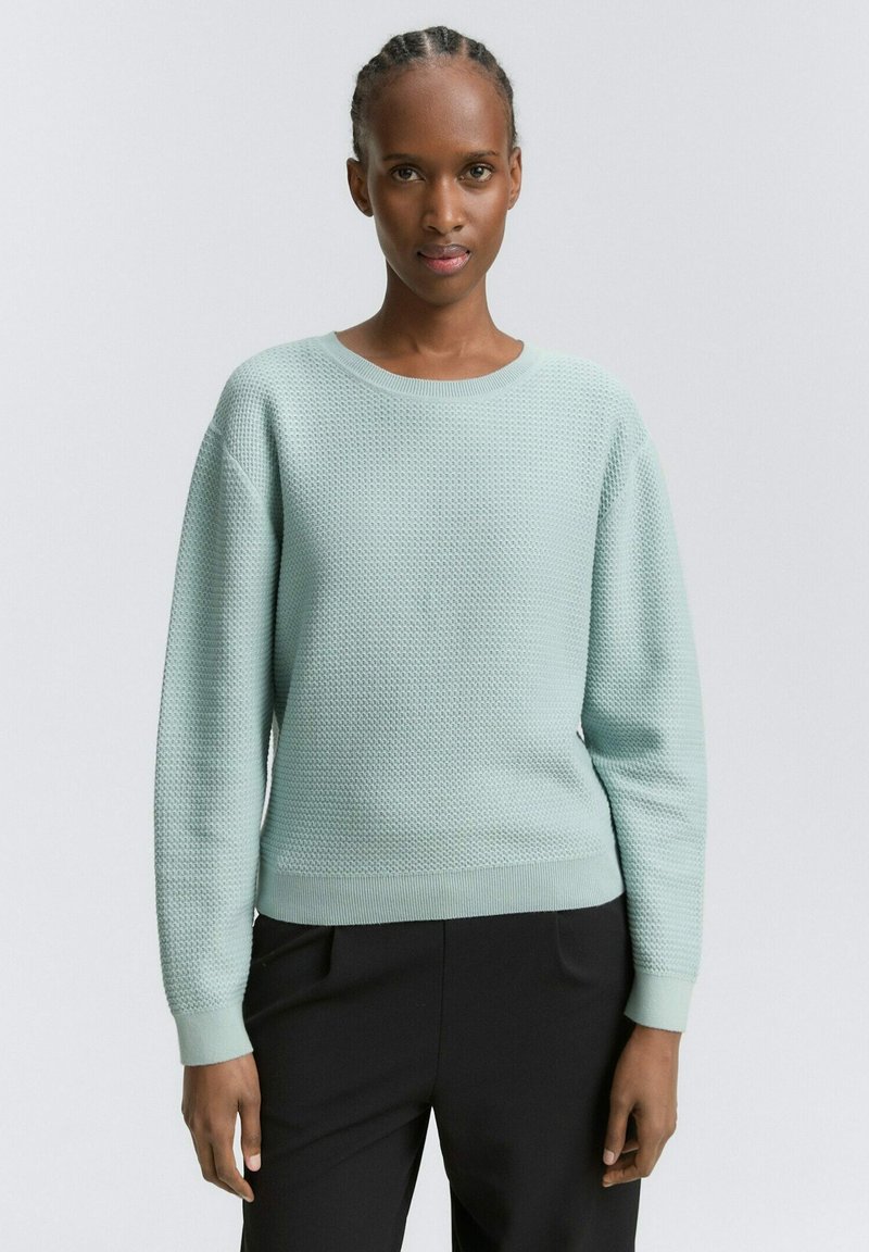 Šviesiai mėtos žalia megztinis su tekstūruota avilio raštu. Turi apvalų iškirpimą, ilgas rankoves ir tvirtinimus bei kraštus su raišteliais.