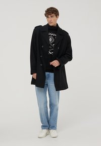 Czarny płaszcz dwurzędowy z wywijanymi klapami, noszony na wzorzystym swetrze, zestawiony z jasnoniebieskimi dżinsami i białymi trampkami.