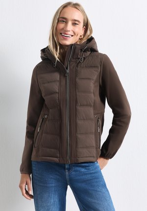 Jeune femme portant une veste matelassée marron à capuche avec des poches zippées et un jean bleu, souriante, une main dans la poche.