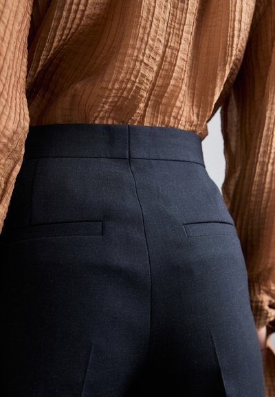 Marineblå højtaljede bukser lavet af struktureret stof, med to baglommer og en strømlinet pasform, parret med en krøllet brun bluse.