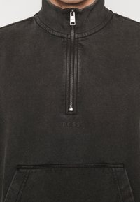 Svart kvartdragets sweatshirt med ribbad krage, framficka och präglad logotyp. Slät textur och avslappnad design. Dragkedjan har ett metalliskt handtag.