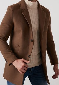 Brun ullblandad kappa med skräddarsydd design, slagkrage, enknäppt front och två svarta knappar. Lagd över en krämfärgad ribbad tröja.