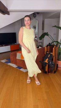 Femme en robe jaune sans manches et sandales blanches debout sur un sol en bois dans un salon avec un canapé marron et un appareil d'exercice.