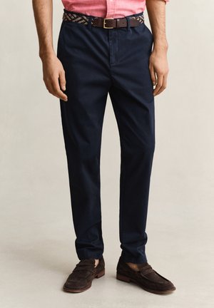 Man som bär mörkblå byxor, brunt läderbälte, bruna mockalofers och en rosa skjorta instoppad, stående mot en ljus bakgrund.