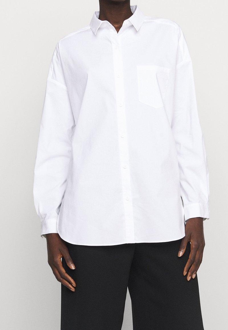 Personne portant une chemise blanche ample à manches longues avec une poche poitrine et un pantalon noir sur un fond uni.