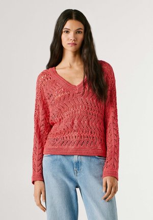 Femme aux longs cheveux foncés portant un pull rouge en maille à col en V et un jean bleu clair, debout devant un fond uni clair.