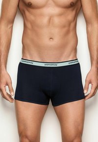 Torso maschile che indossa slip blu navy con cintura grigia chiara sul quale è presente il logo "yamamay", in piedi contro uno sfondo neutro.